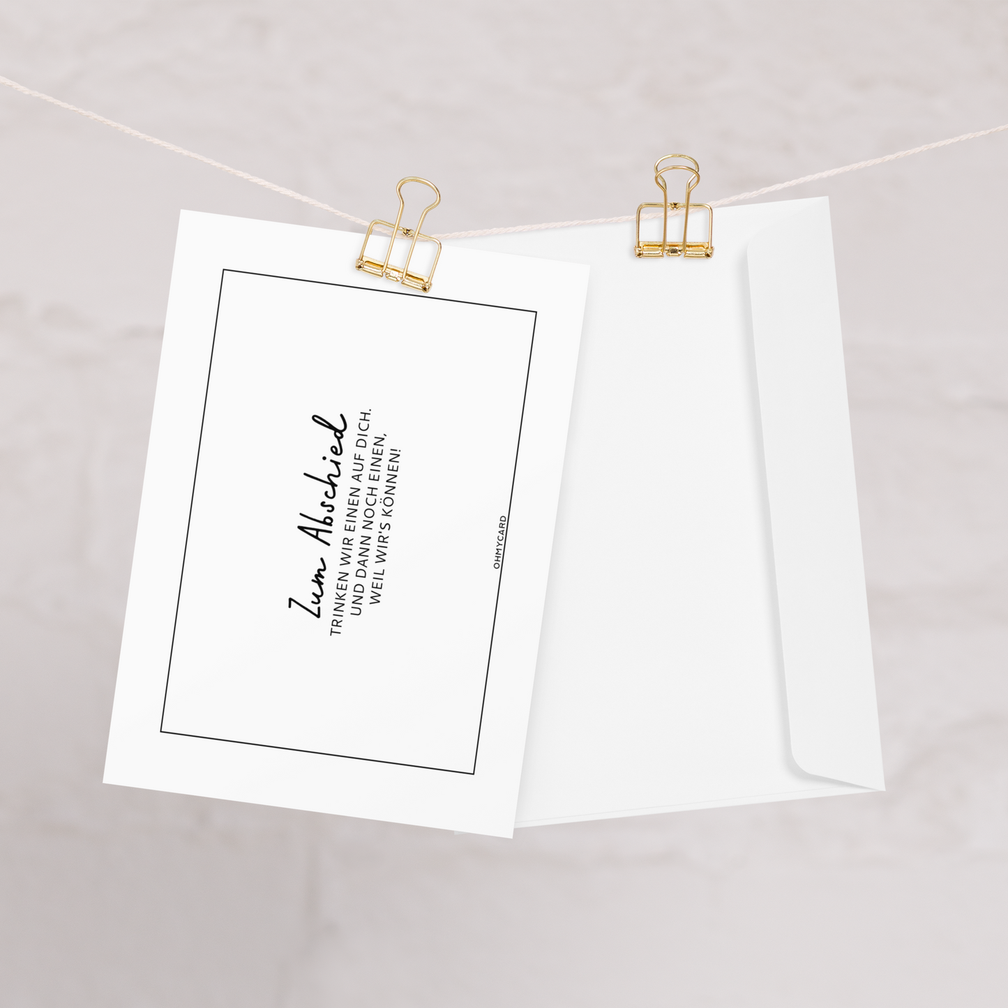 Eine Glückwunschkarte von ohmycard mit dem Text "Zum Abschied!" und passendem Umschlag wird mit goldenen Binderclips an einer Schnur befestigt.