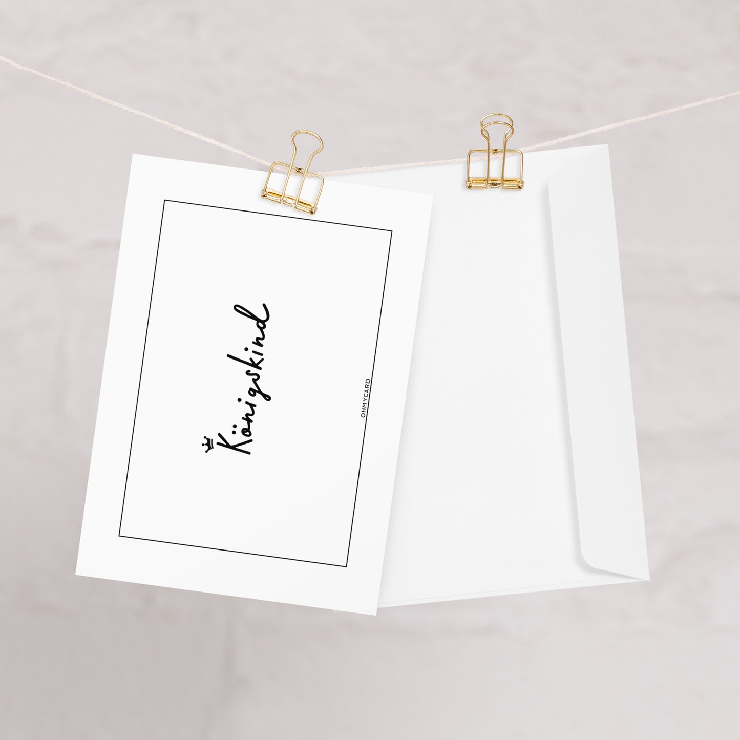 Die weiße Königskind-Karte von ohmycard mit kleiner Krone und ermutigender Botschaft hängt an einer goldenen Klammer neben passendem Umschlag - ein liebevolles Geschenk für besondere Anlässe.
