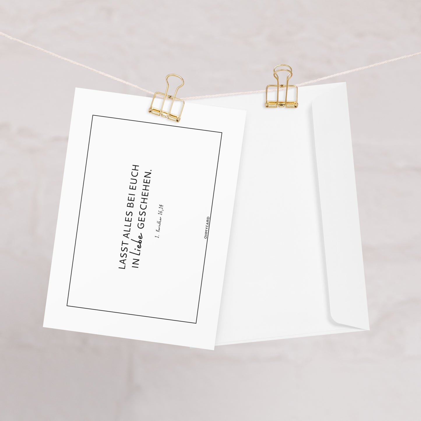 Die Hochzeitskarte "Lasst alles in Liebe geschehen" von ohmycard mit Bibelvers auf Deutsch hängt an einer Schnur neben dem passenden weißen Umschlag, beide mit goldenen Binderclips befestigt. Der Hintergrund ist hell und minimalistisch.