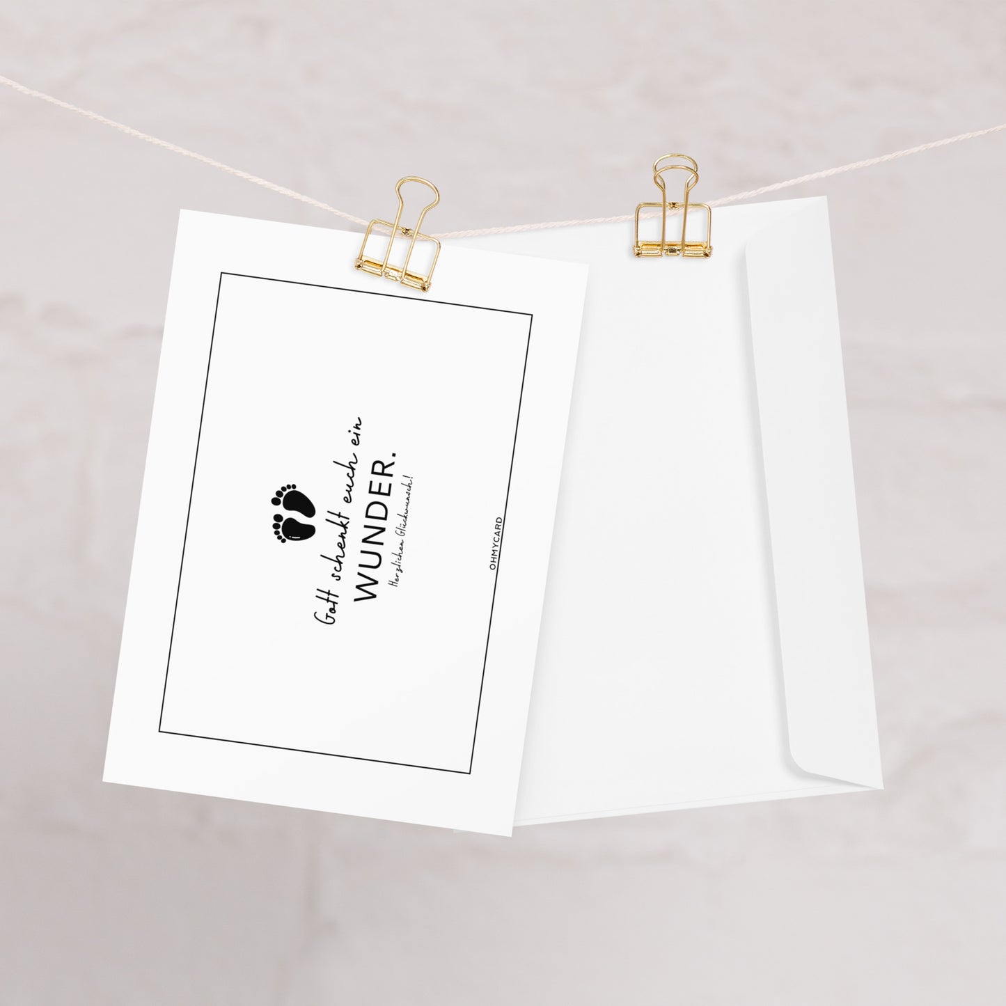 Die ohmycard Babykarte "Gott schenkt euch ein Wunder" mit Umschlag hängt an einer Schnur mit goldenen Clips. Sie zeigt kleine Fußabdrücke und den Text "Gott schenkt euch ein Wunder" auf hellem Hintergrund.