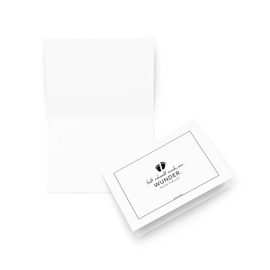 Eine aufgeschlagene weiße "Gott schenkt euch ein Wunder"-Karte von ohmycard ist neben ihrem Umschlag abgebildet, der schwarze Schrift und kleine Fußabdrücke über den Worten WUNDER und Glück ist das, was du daraus machst zeigt.