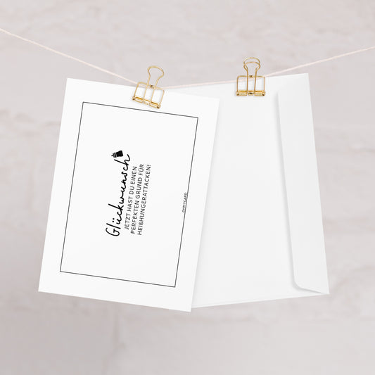 Eine weiße ohmycard "Heißhunger" Glückwunschkarte Schwangerschaft mit deutschem Text und einer kleinen Abschlusskappe ist mit goldenen Büroklammern an einer Schnur befestigt. Daneben hängt ein Briefumschlag vor einem weichen, hellen Hintergrund.