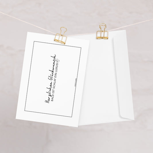Zwei weiße "Bald ist Schlaf ein Luxus"-Karten von ohmycard hängen an einer Schnur mit goldenen Klammern; eine zeigt die deutsche Vorderseite, die andere die Rückseite. Der sanft verschwommene, neutrale Hintergrund setzt einen eleganten Akzent.