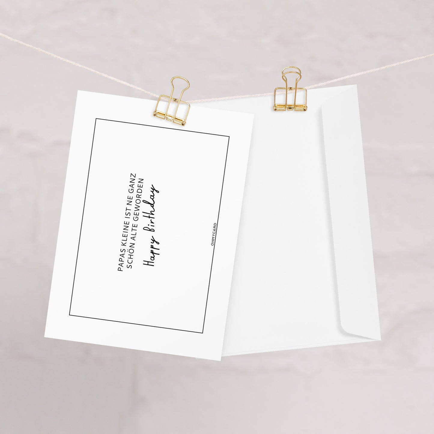 Die Geburtstagskarte "Papas KLEINE" von ohmycard mit deutschem Text und Happy Birthday in Schreibschrift hängt an einer Schnur mit goldenen Klammern neben einem schlichten weißen Umschlag, alles vor einem sanft verschwommenen Hintergrund.