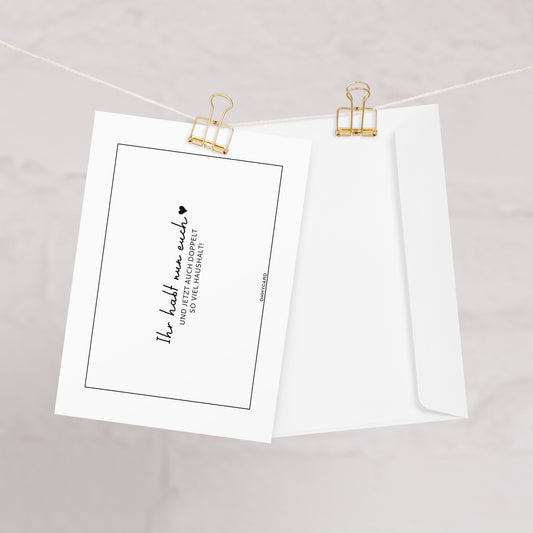 Eine weiße "Doppelt so viel Haushalt"-Karte von ohmycard mit schwarzem Text wird neben einem passenden Umschlag mit goldenen Büroklammern an einer Schnur befestigt - eine perfekte humorvolle Glückwunschkarte zur Hochzeit.