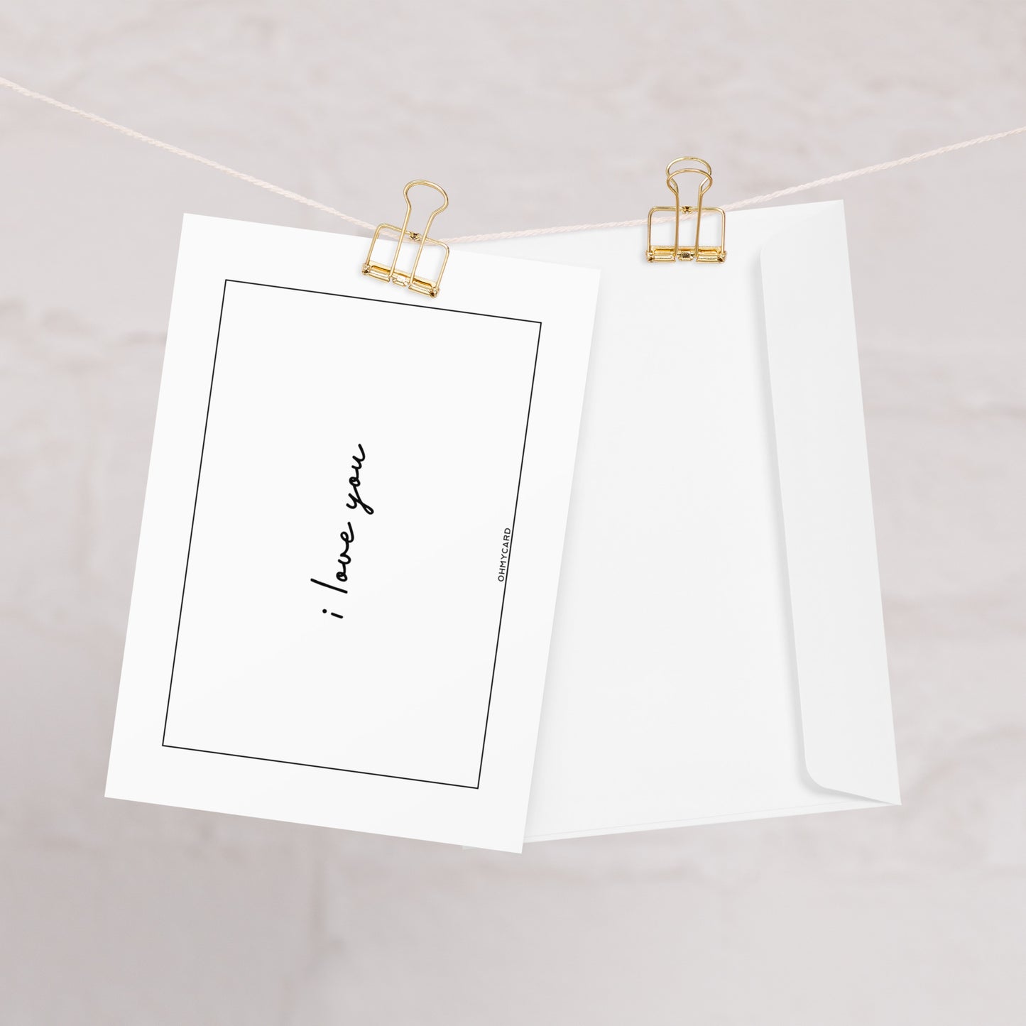Eine weiße "I love you" Valentinstagskarte von ohmycard ist neben einem weißen Briefumschlag an einer Schnur befestigt, beide werden von goldenen Büroklammern vor einem sanft verschwommenen Hintergrund gehalten.