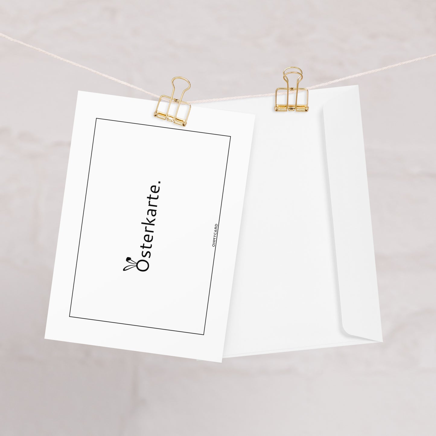 Die ohmycard Osterkarte ist eine weiße Grußkarte mit schwarzem Schriftzug "Osterkarte", die mit einer goldenen Büroklammer an einer Schnur befestigt ist, daneben ein schlichter weißer Umschlag vor hellem Hintergrund - ideal für frohe Ostern-Wünsche.