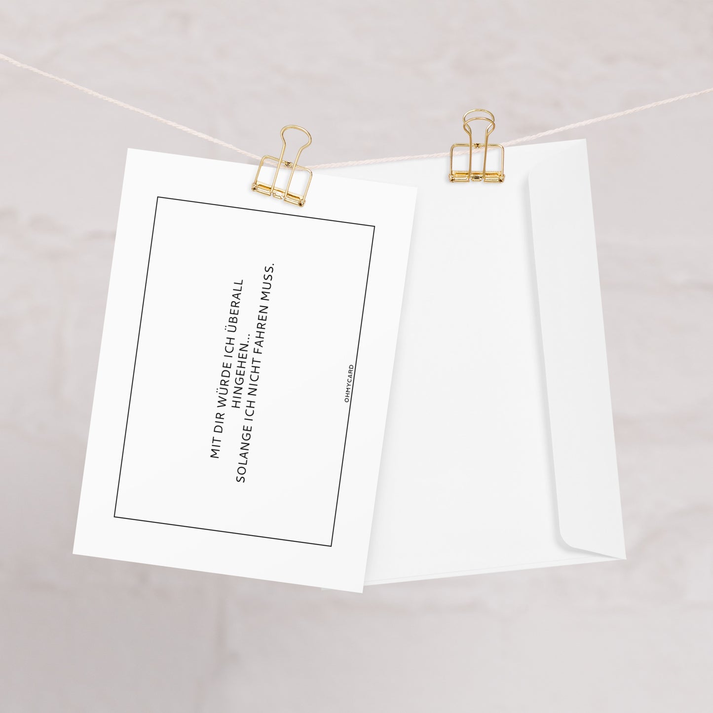 Die humorvolle Karte "Mit dir würde ich überall hingehen" von ohmycard mit deutscher Aufschrift hängt mit goldenem Binder Clip an einer Schnur. Daneben liegt eine schlichte weiße Hülle vor hellem Hintergrund.