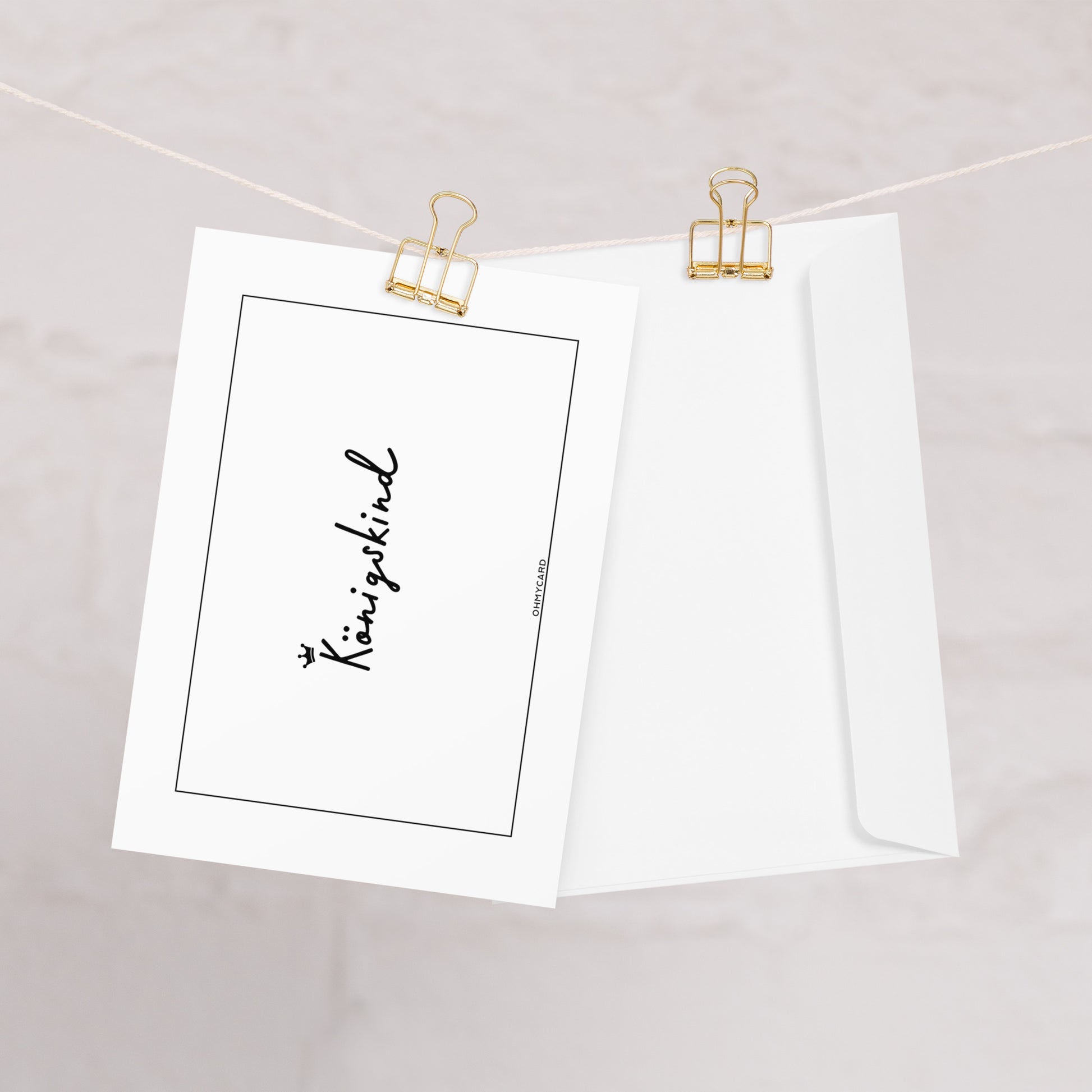 Die weiße Königskind-Karte von ohmycard mit kleiner Krone und ermutigender Botschaft hängt an einer goldenen Klammer neben passendem Umschlag - ein liebevolles Geschenk für besondere Anlässe.