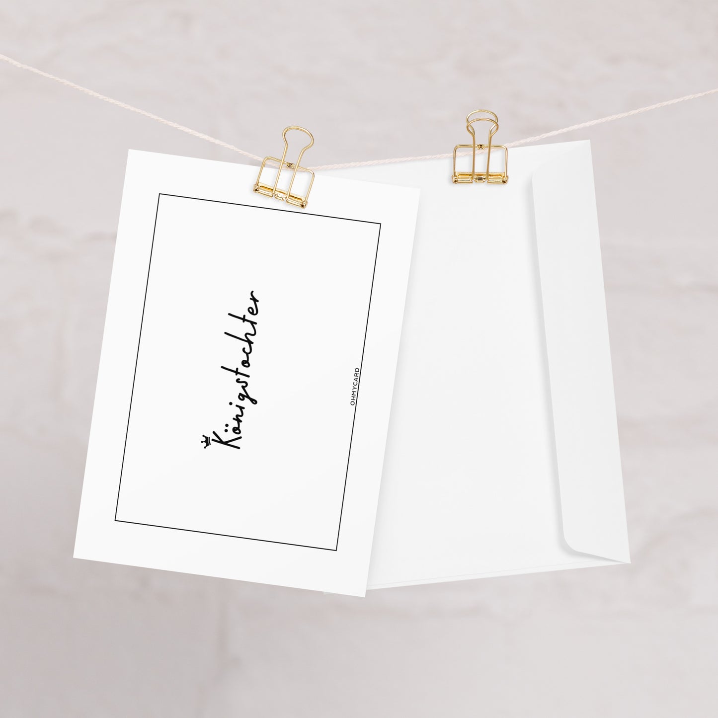 Eine weiße ohmycard "Königstocher"-Karte mit schwarzer Handschrift "Königstöchter" hängt an goldenen Heftklammern neben einem weißen Umschlag - ideal als Ermutigungskarte Frau, vor einem hellen, neutralen Hintergrund.