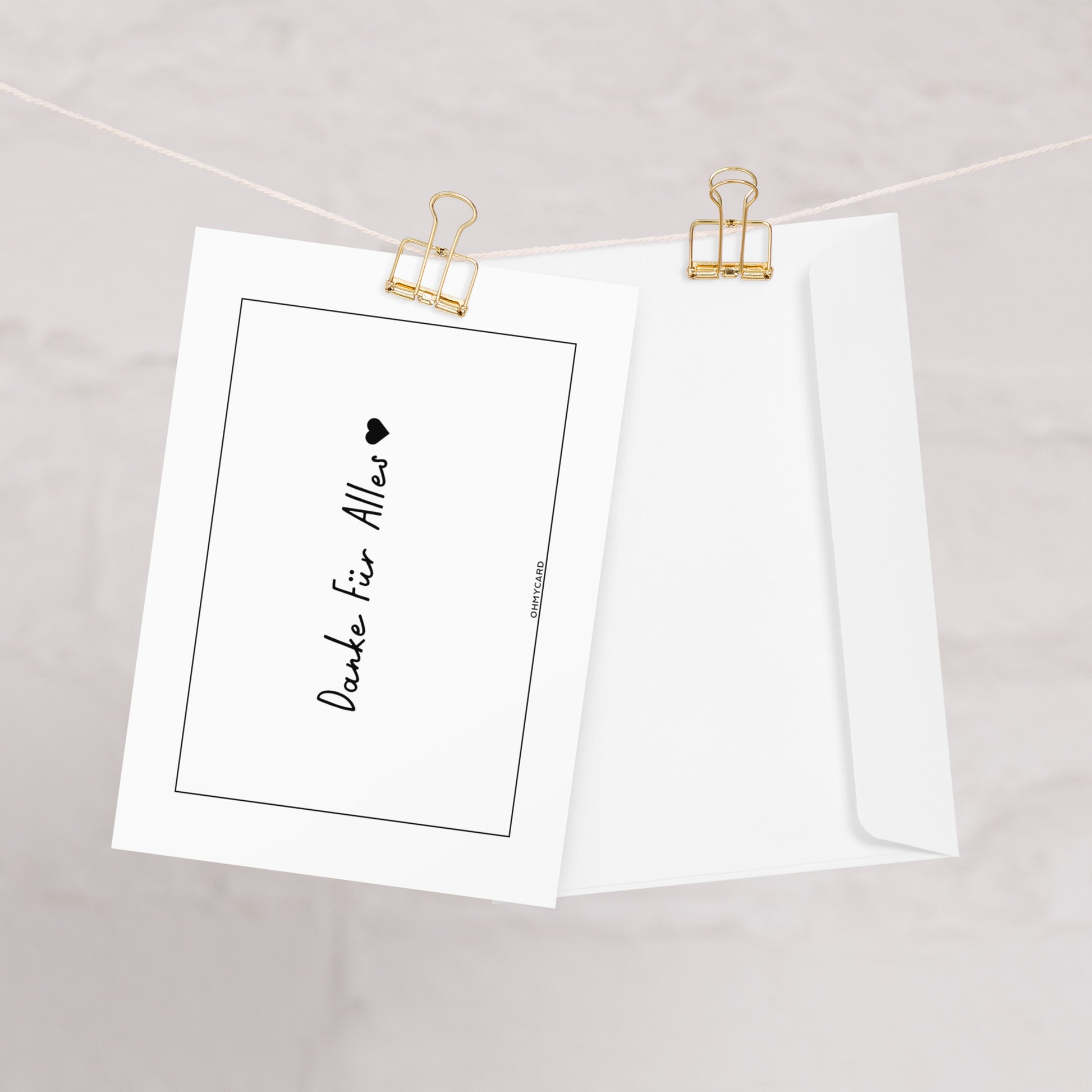 Die ohmycard Danke Für Alles HERZ: eine weiße Karte mit "Danke für Alles" und einem Herz, die zusammen mit dem passenden Umschlag mit goldenen Büroklammern an einer Schnur befestigt ist, vor einem hellen Hintergrund.