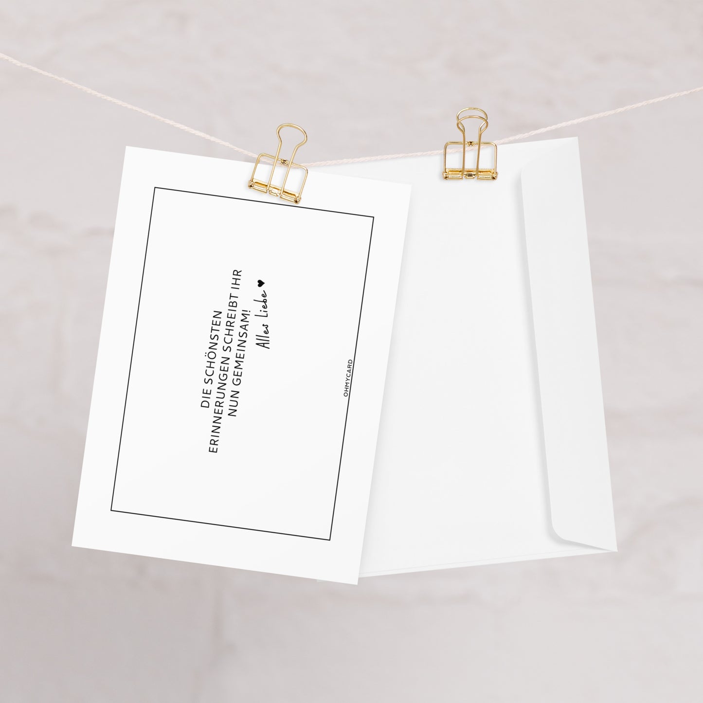 Die Karte "Erinnerungen schreiben" von ohmycard mit deutschem Text und Unterschrift ist neben einem schlichten Briefumschlag an einer weißen Schnur mit goldenen Heftklammern vor hellem Hintergrund befestigt.