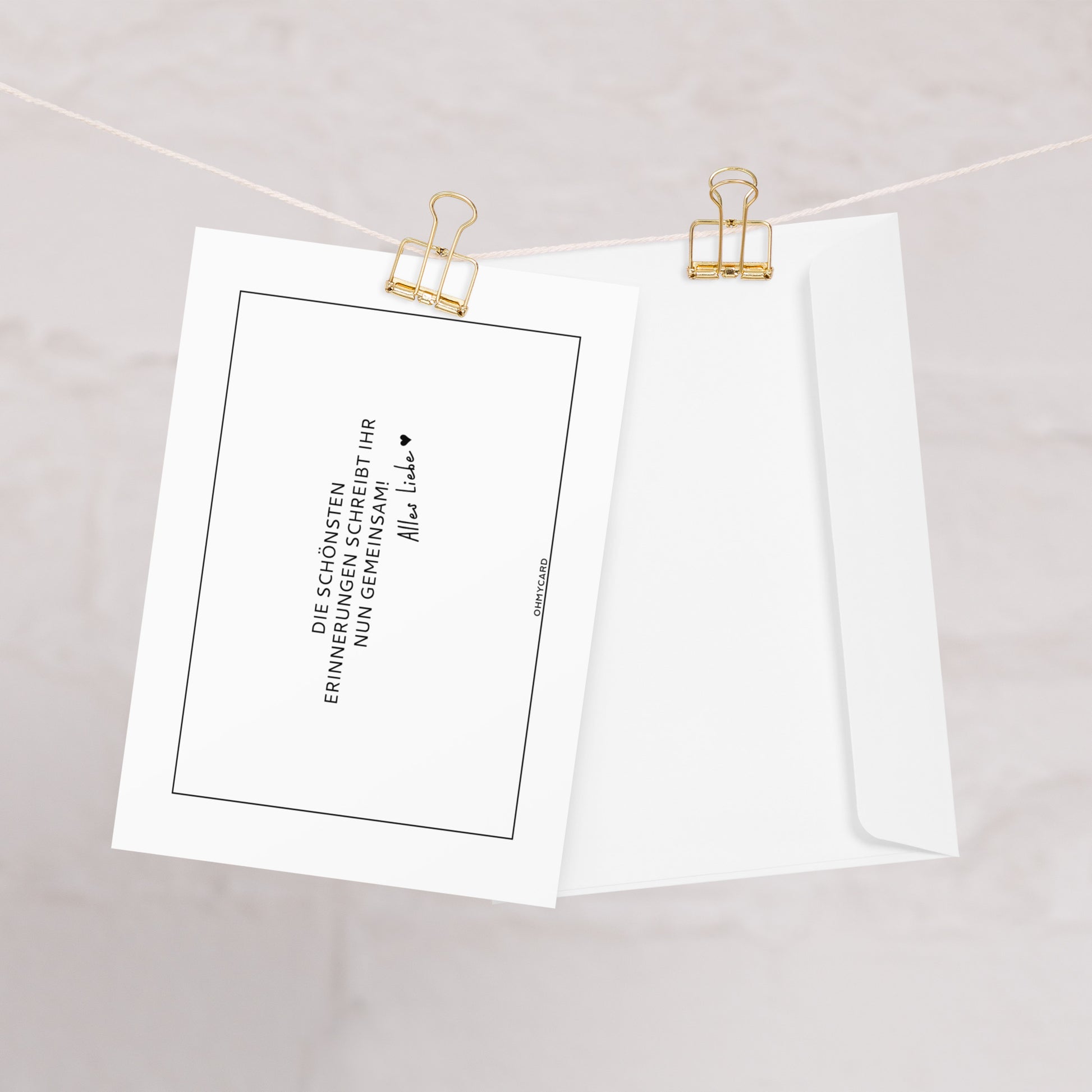 Die Karte "Erinnerungen schreiben" von ohmycard mit deutschem Text und Unterschrift ist neben einem schlichten Briefumschlag an einer weißen Schnur mit goldenen Heftklammern vor hellem Hintergrund befestigt.