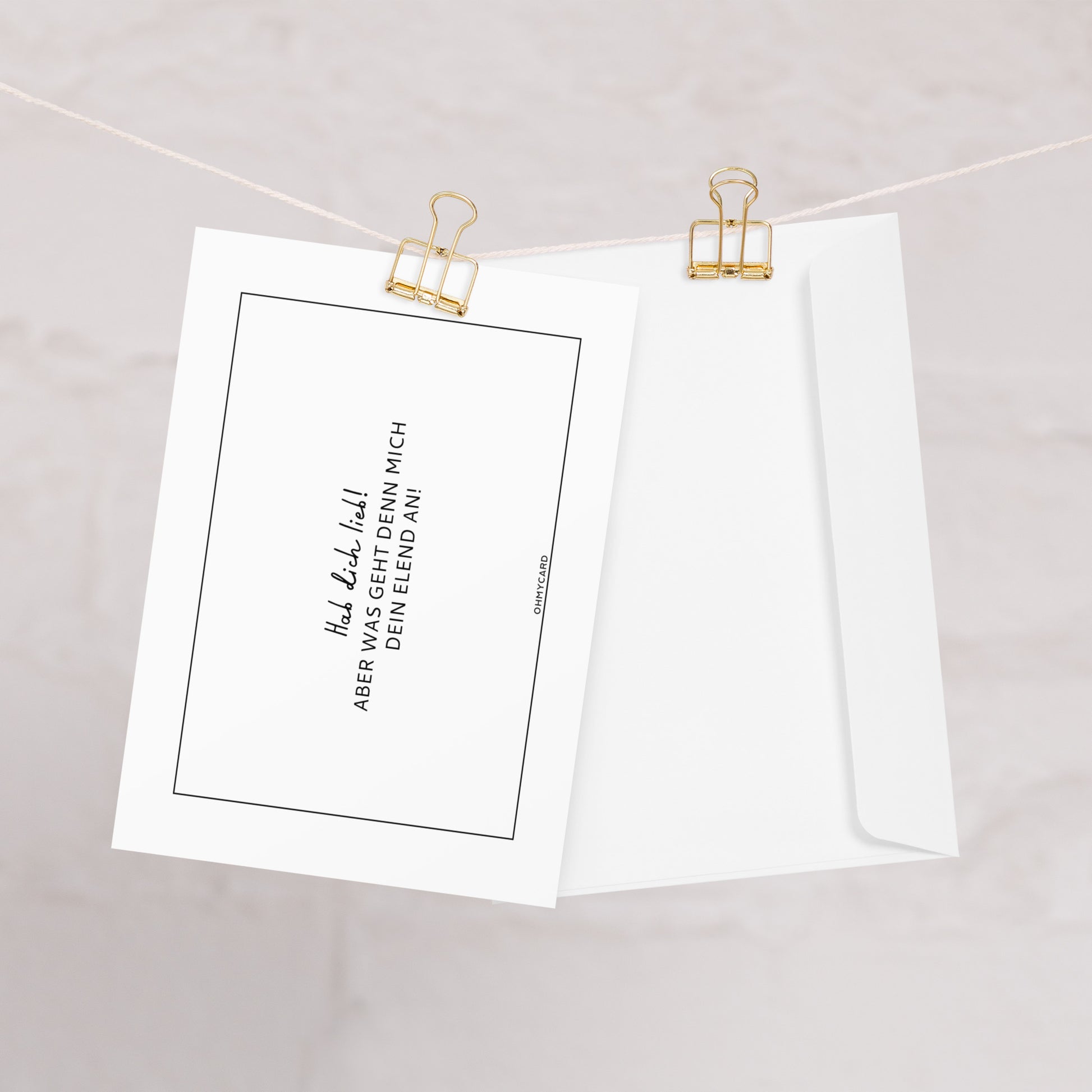 Die weiße Grußkarte "Was geht mich dein Elend an!" von ohmycard mit deutschem Sarkasmus und schwarzem Humor hängt samt weißem Umschlag an goldenen Clips vor hellem Hintergrund.